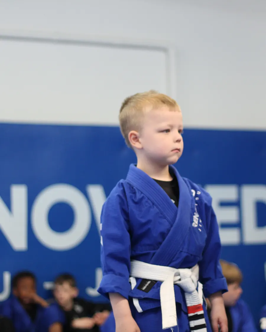 A young child in a blue gi standing at the edge of the mat, watching the class