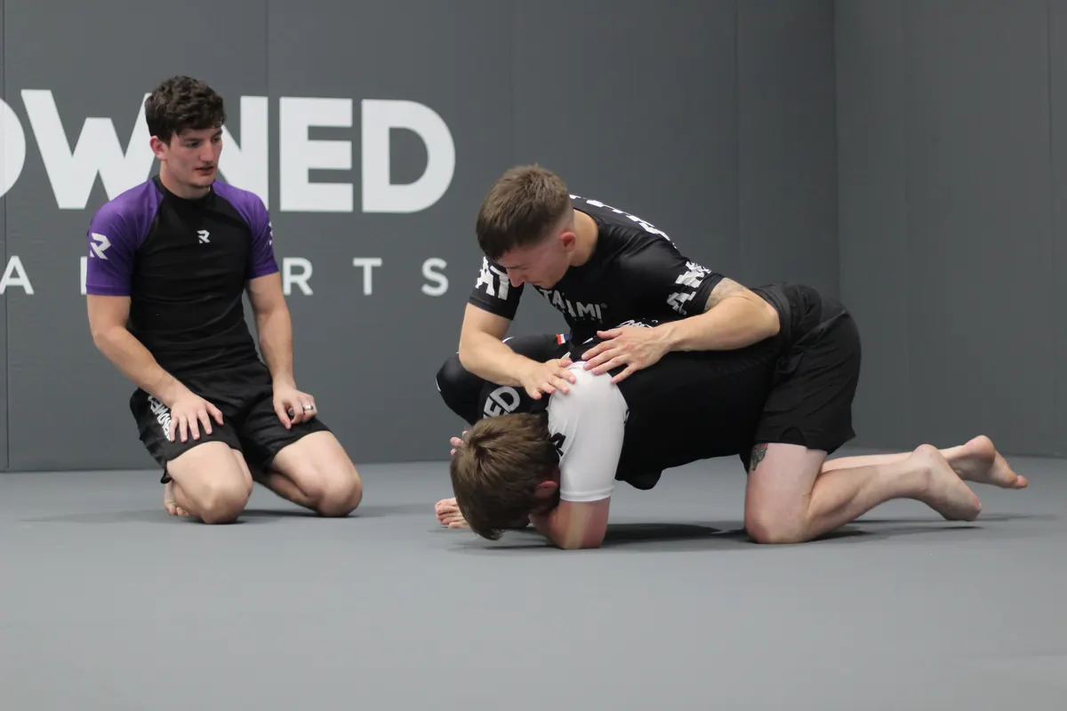 Several pairs of adults training at once during a no-gi class, a coach kneeling beside one of them